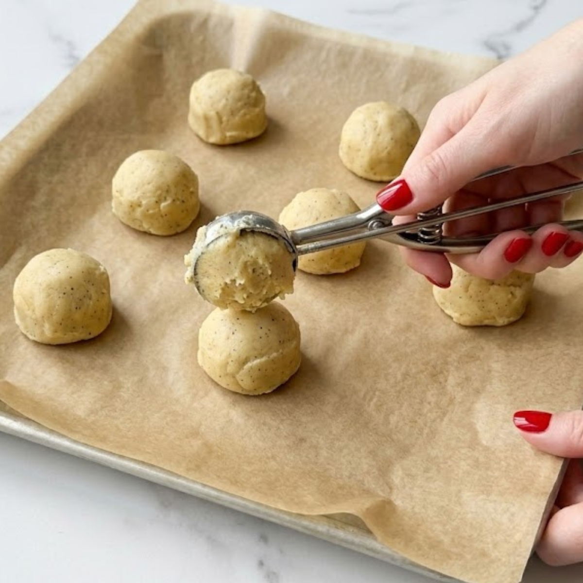 Close-up of a young woman's hands with classic red nails using a cookie scoop to place uniform, round balls of dense cookie dough onto a parchment-lined baking sheet on a white marble counter.