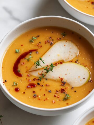 A speckled white bowl of smooth golden spicy apple soup garnished with thin apple slices, fresh thyme, red chili flakes, and a drizzle of spiced oil, on a marble surface with additional bowls and thyme sprigs nearby.
