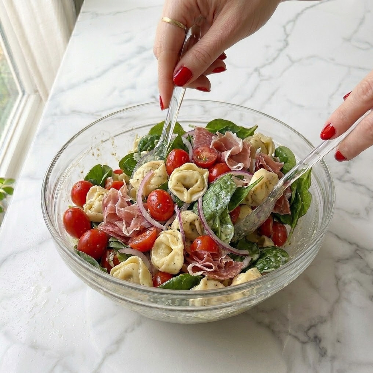 Close-up dyal young woman's hands with classic red nails gently tossing cooled cheese tortellini pasta with halved cherry tomatoes, red onion slices, and cured meat ribbons f a large bowl on a white marble counter, matching