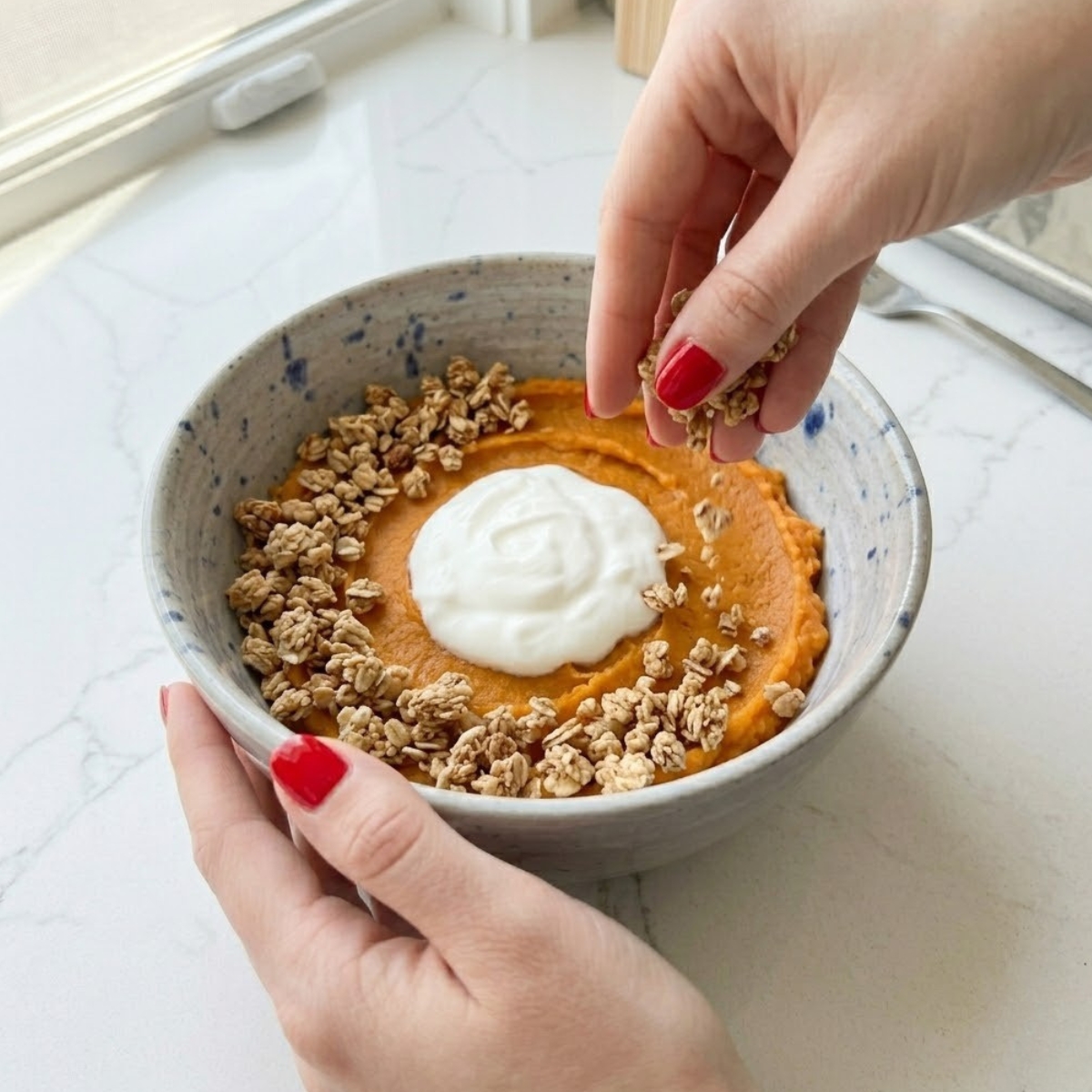 Close-up view of a young woman's hand with classic red nails scattering crunchy granola in an arc around a dollop of Greek yogurt resting on a smooth sweet potato base inside a speckled serving bowl.