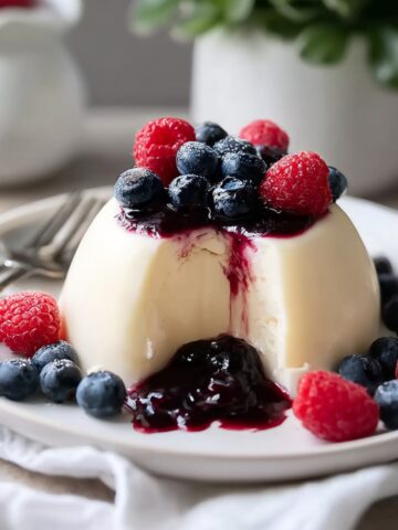 A domed Bavarian cream unmolded on a white plate, topped with dark berry compote and fresh blueberries and raspberries, with a spoonful taken out revealing the silky cream interior, surrounded by scattered fresh berries and a fork on the side.