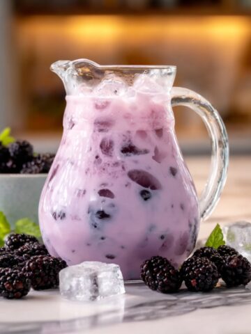 A glass pitcher of blackberry coconut pink drink with a soft purple hue and whole blackberries floating throughout, served over ice, surrounded by fresh blackberries, ice cubes, and mint sprigs on a marble surface with a warm kitchen background.