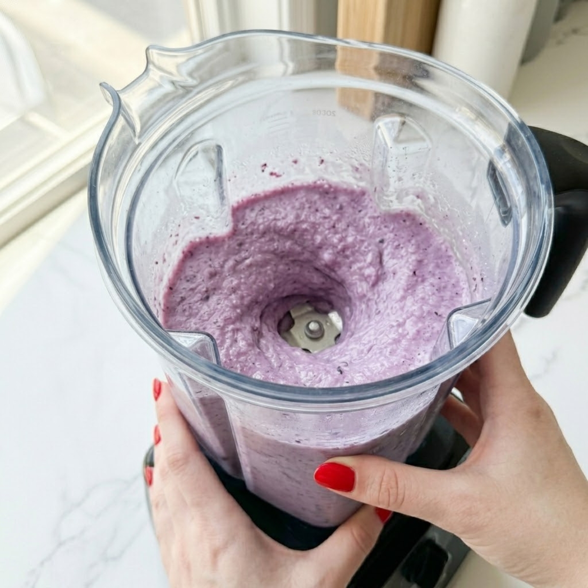 Close-up view of a young woman's hand with classic red nails operating a blender, with a thick, slushy, vibrant purple blueberry colada mixture actively swirling inside the frosted pitcher.