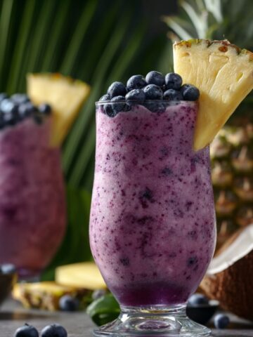 Two hurricane glasses of frozen blueberry colada with a vibrant purple blended base, topped with fresh blueberries and a pineapple wedge, surrounded by scattered blueberries, pineapple chunks, a coconut half, and tropical palm leaves in the background.