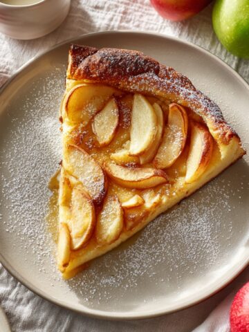 A slice of German apple pancake on a white plate, topped with caramelized apple slices and dusted with powdered sugar, surrounded by fresh red and green apples on a linen cloth.