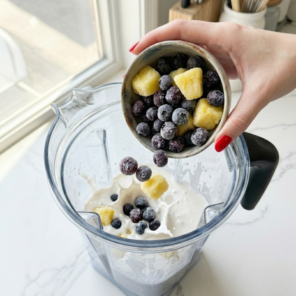 Close-up view of a young woman's hand with classic red nails dropping frozen blueberries and pineapple chunks into a blender pitcher filled with white coconut cream on a white marble counter.