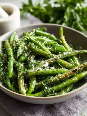 A ceramic bowl of oven roasted green beans with slightly charred edges, sprinkled with grated Parmesan cheese and fresh herbs, with a white mortar and pestle and fresh herbs in the background.