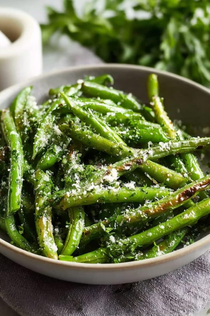 A ceramic bowl of oven roasted green beans with slightly charred edges, sprinkled with grated Parmesan cheese and fresh herbs, with a white mortar and pestle and fresh herbs in the background.