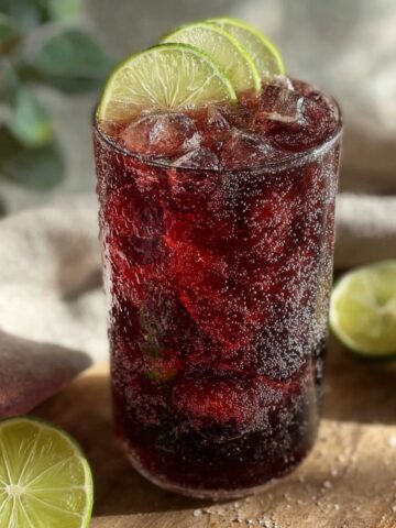 A tall glass of deep ruby hibiscus ginger iced tea over ice, garnished with lime slices on the rim, surrounded by halved limes on a wooden surface with a linen cloth and greenery in the background.
