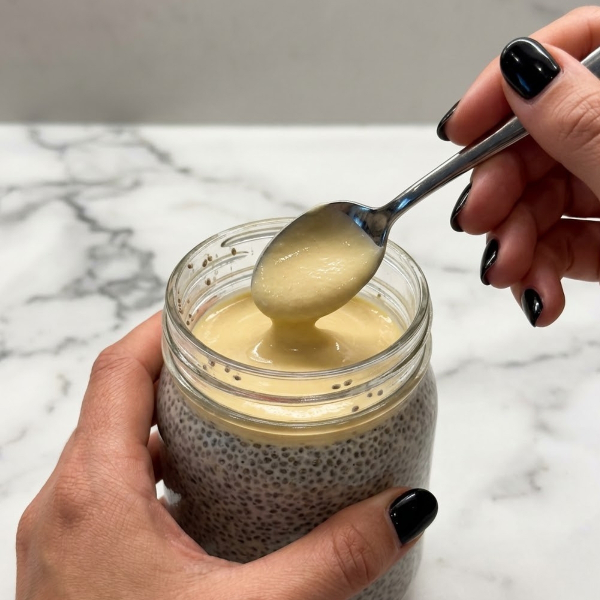 Close up view of a woman's hands with glossy black nails using a spoon to layer thick pale yellow lemon cream over a set chia pudding base inside a glass jar.