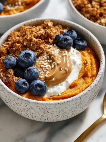 A speckled ceramic bowl of smooth sweet potato puree topped with yogurt, a swirl of peanut butter, granola, fresh blueberries, and sesame seeds, with a gold spoon alongside on a marble surface.