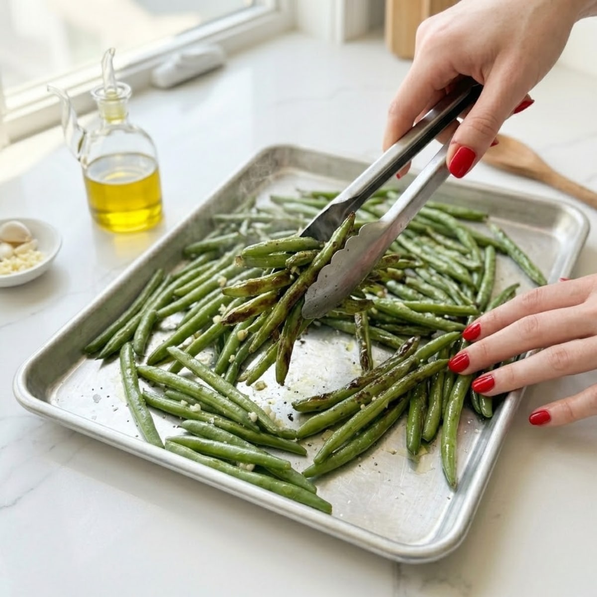 Close-up view of a young woman's hands with classic red nails using tongs to toss hot, tender-crisp green beans with caramelized brown spots on a baking sheet.