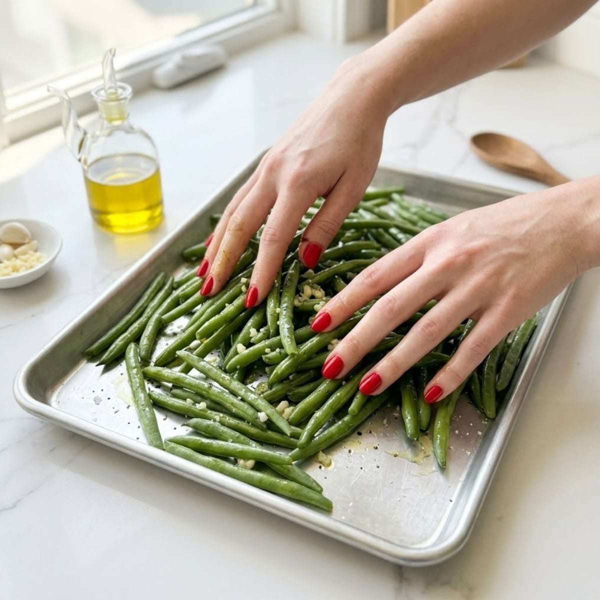 Close-up view of a young woman's hands with classic red nails tossing fresh green beans with olive oil and minced garlic on a large rimmed baking sheet on a white marble counter.