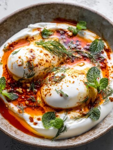 Overhead view of Turkish eggs (çılbır) in a rustic ceramic bowl, with two poached eggs nestled on a bed of creamy white yogurt, drizzled with red chili butter, and garnished with fresh dill and mint leaves.