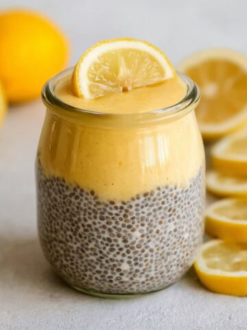 "A glass jar of Vegan Lemon Cream Pie Chia Pudding layered with chia pudding and creamy lemon curd, topped with a fresh lemon slice, surrounded by sliced lemons."