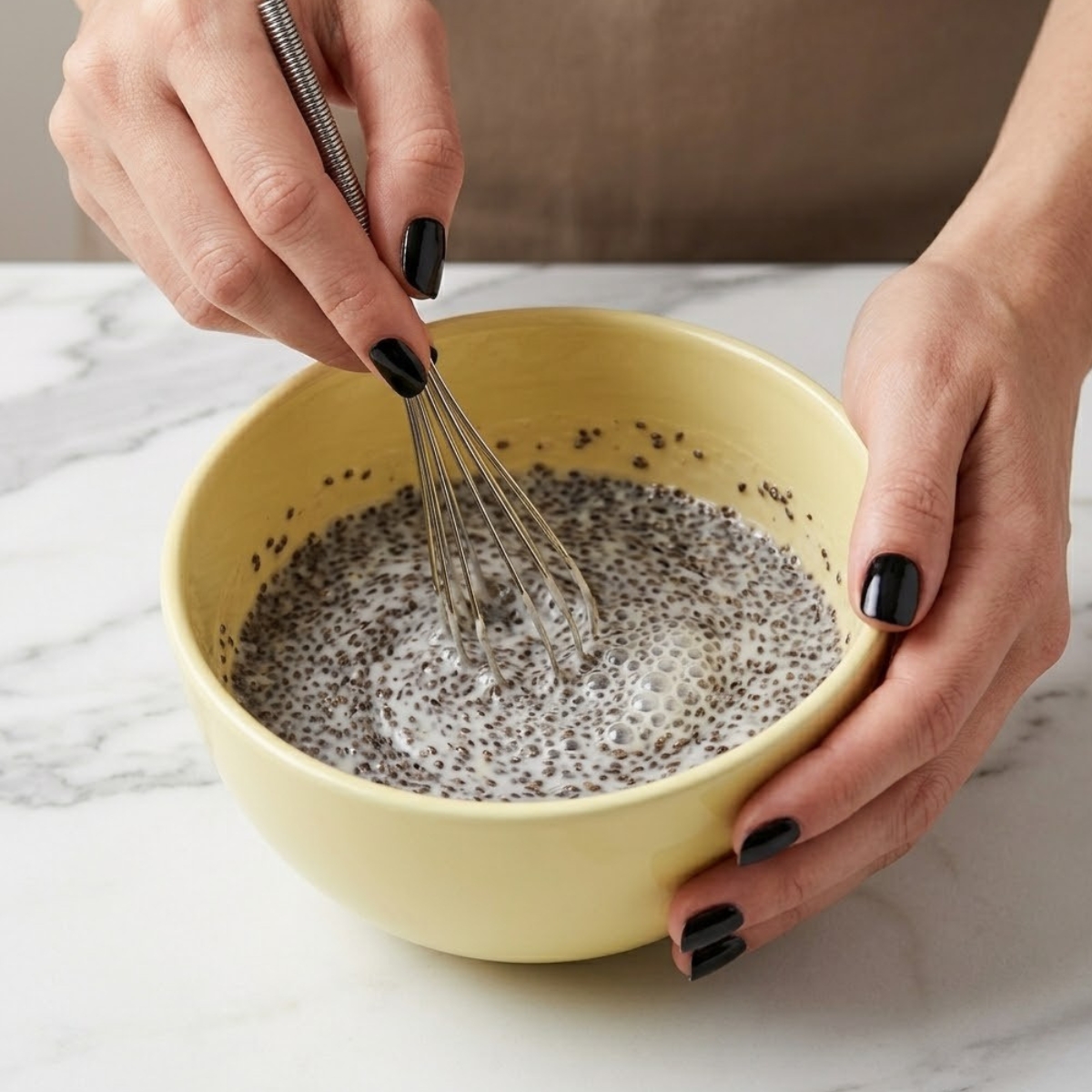 Close up view of a woman's hands with glossy black nails whisking chia seeds and coconut milk in a pastel yellow mixing bowl on a marble counter.