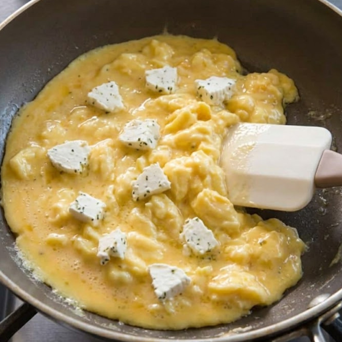 A close-up shot of a silicone spatula (referenced from image_12.png) gently drawing through the golden egg mixture in a non-stick skillet. Large, soft, glossy curds (visible texture from image_13.png) are forming, and the white, herb-flecked Boursin cheese (ingredient seen in image_12.png) is scattered across the surface, beginning to melt.