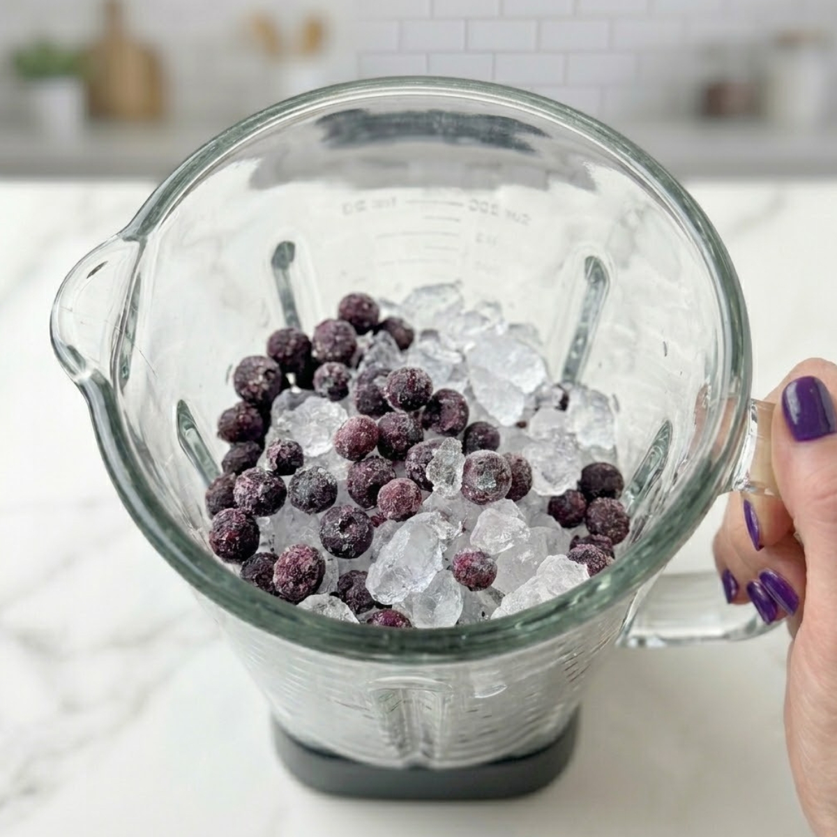 Extreme close-up of hands with deep purple nails pouring frozen blueberries and crushed ice into a blender.