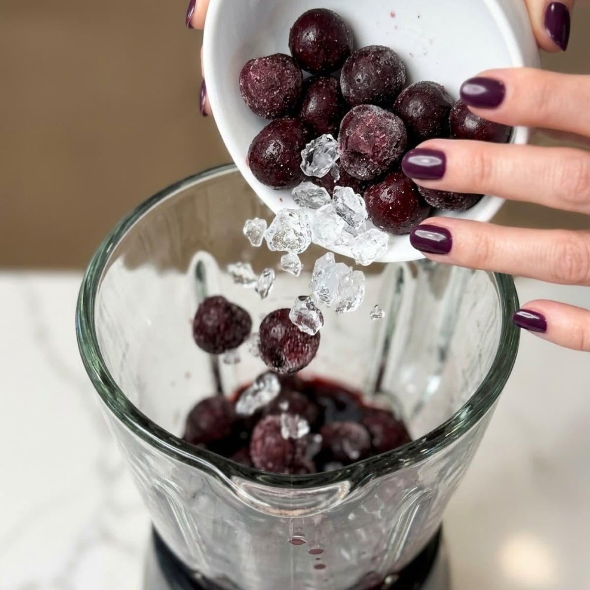 Extreme close-up of hands with plum nails adding frozen cherries and ice to a blender.