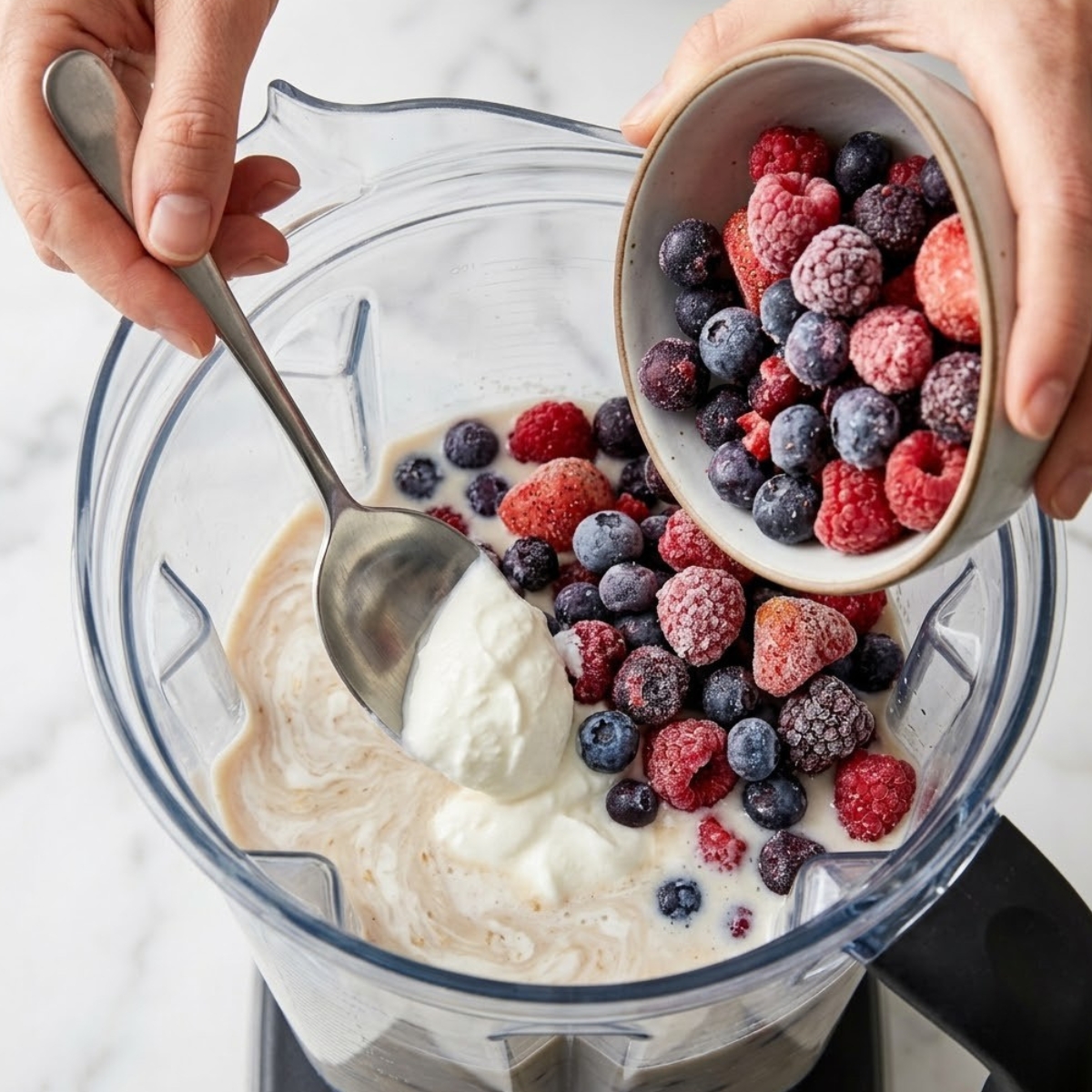 A close-up view capturing hands actively loading the blender jar. A spoon is layering full-fat Greek yogurt over the milk, and frozen berries are being poured from a bowl, referencing the specific ingredients and textures from image_32.png.