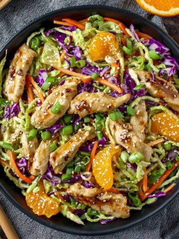 Overhead view of a vibrant Asian chicken salad in a dark ceramic bowl, filled with shredded green cabbage, purple cabbage, julienned carrots, sliced green onions, and mandarin orange segments, topped with glazed teriyaki chicken strips sprinkled with sesame seeds, placed on a gray textured linen with wooden salad tongs alongside and halved fresh oranges in the upper corner.