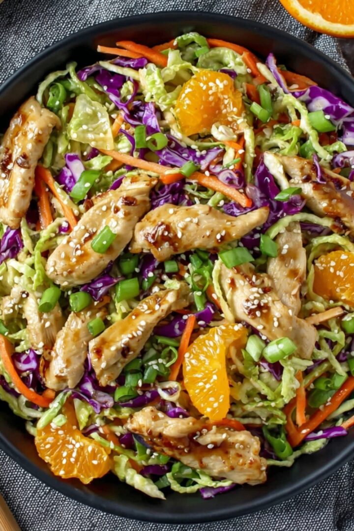 Overhead view of a vibrant Asian chicken salad in a dark ceramic bowl, filled with shredded green cabbage, purple cabbage, julienned carrots, sliced green onions, and mandarin orange segments, topped with glazed teriyaki chicken strips sprinkled with sesame seeds, placed on a gray textured linen with wooden salad tongs alongside and halved fresh oranges in the upper corner.