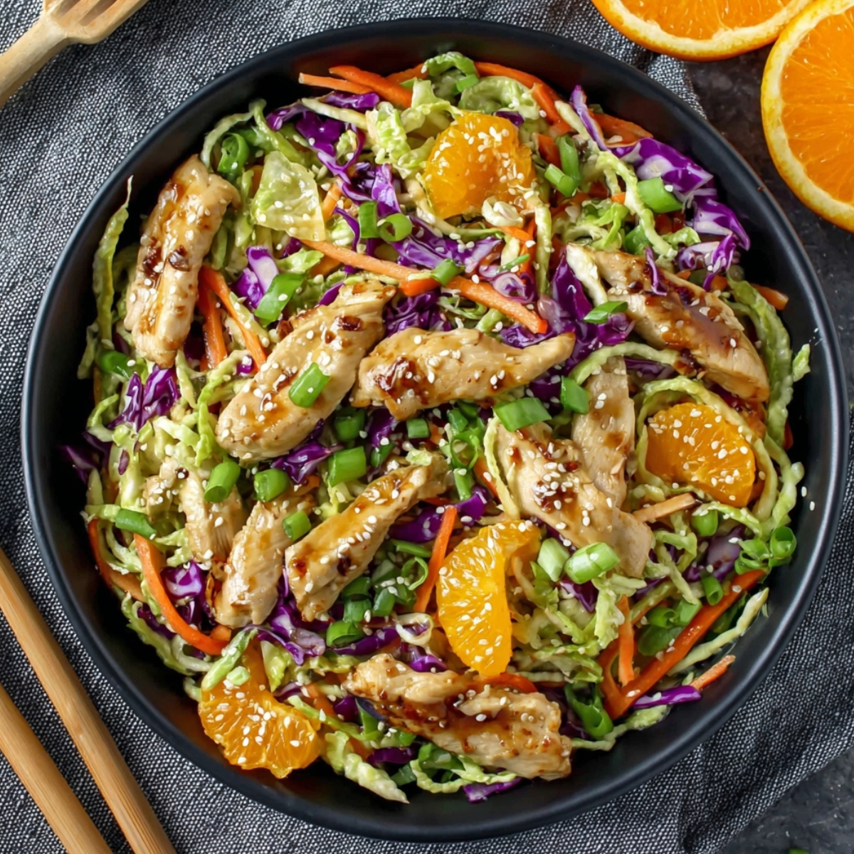 Overhead view of a vibrant Asian chicken salad in a dark ceramic bowl, filled with shredded green cabbage, purple cabbage, julienned carrots, sliced green onions, and mandarin orange segments, topped with glazed teriyaki chicken strips sprinkled with sesame seeds, placed on a gray textured linen with wooden salad tongs alongside and halved fresh oranges in the upper corner.