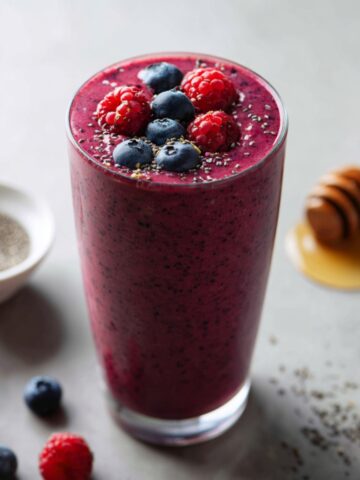 A tall clear glass filled with a deep crimson berry probiotic smoothie speckled with chia seeds throughout, topped with fresh blueberries, raspberries, and a sprinkle of chia seeds, surrounded by scattered fresh blueberries and raspberries on a light gray surface, with a small white bowl of chia seeds and a wooden honey dipper resting in a pool of golden honey alongside.