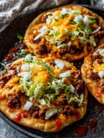 An aerial close-up shot of three open-faced "Big Mac Tacos" arranged in a large black skillet. The tacos feature thick, golden-brown fried flatbreads topped with seasoned ground beef, melted cheddar cheese, shredded lettuce, and diced white onions, with dollops of reddish-orange burger sauce scattered across the pan.