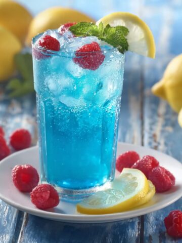 A tall clear glass filled with a vibrant electric blue raspberry lemonade packed with ice and fresh raspberries, garnished with a lemon wedge on the rim and a fresh mint sprig, served on a white plate surrounded by fresh raspberries and lemon slices, on a weathered light blue wooden surface with whole lemons and a halved lemon softly blurred in the sunny background.