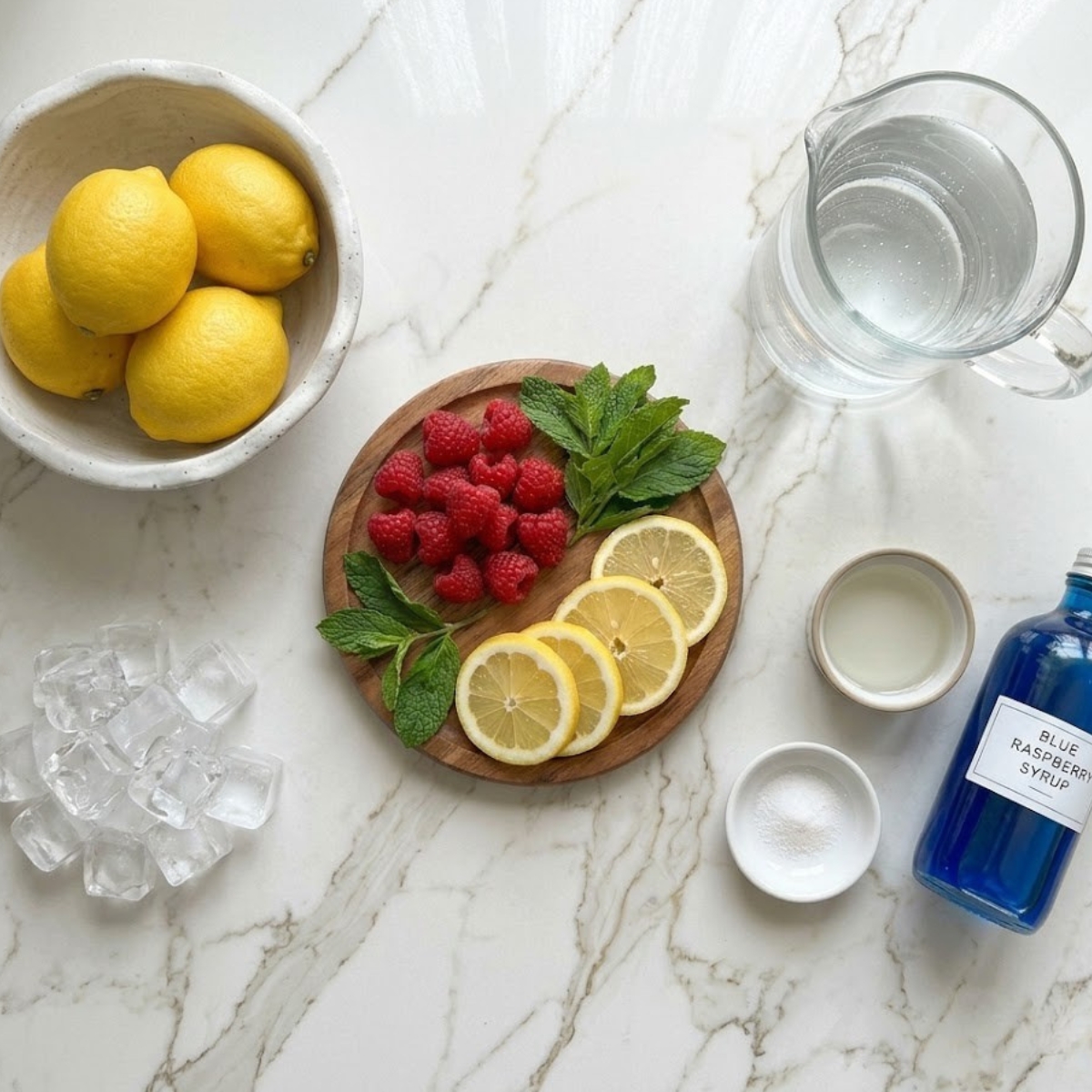 A high-angle flat-lay photograph capturing all raw ingredients spread across a light marble kitchen counter. It includes a bowl of fresh lemons, a pitcher of cold water, a bottle of bright blue raspberry syrup, simple syrup, a pinch of salt, ice cubes, fresh raspberries, lemon slices, and mint sprigs, matching the clean aesthetic of the finished drink image.