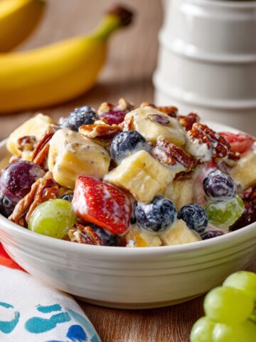 A creamy breakfast fruit salad in a white bowl featuring banana slices, blueberries, strawberries, green and red grapes, and pecans tossed in a yogurt dressing, with fresh bananas and grapes in the background.
