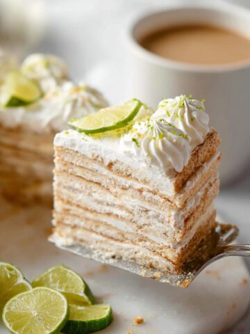 A slice of Carlota de Limón being lifted on a silver cake server, revealing beautiful distinct layers of golden Maria cookies alternating with creamy lime filling, topped with swirled whipped cream rosettes, lime zest, and a fresh lime wheel, with sliced limes scattered on a white marble surface, an additional slice and a cup of coffee softly blurred in the background.