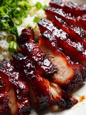 Close-up of thickly sliced char siu chicken with a deeply lacquered, glossy mahogany-red glaze and charred edges, fanned out over steamed white rice, revealing a juicy pale interior with a vivid red smoke ring beneath the caramelized crust, garnished with sliced green onions and fresh herbs on a white plate.