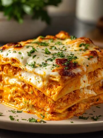 A towering square slice of cheesy buffalo ranch chicken lasagna on a beige ceramic plate, showing multiple distinct layers of pasta sheets alternating with vibrant orange buffalo ranch chicken filling and creamy white cheese sauce, topped with a golden bubbly melted mozzarella crust garnished with chopped fresh parsley, with a bowl of fresh herbs and a white ceramic container softly blurred in the background.