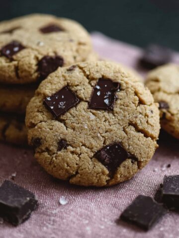 Close-up of three golden-brown chocolate chip protein cookies studded with large dark chocolate chunks and sprinkled with flaky sea salt, arranged on a dusty pink linen with extra dark chocolate pieces scattered around, against a dark green background.