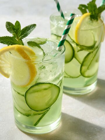 Two clear glass tumblers filled with refreshing pale green cucumber lemon mint infused water, packed with ice and thin cucumber slices, each garnished with a lemon wheel on the rim, a fresh mint sprig, and a green and white striped paper straw, set on a light gray surface with soft natural shadows.