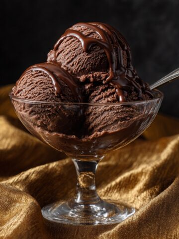 Three scoops of rich dark chocolate sorbet in a glass dessert coupe, drizzled with chocolate sauce and served with a vintage silver spoon, on a luxurious gold satin fabric against a dark background.