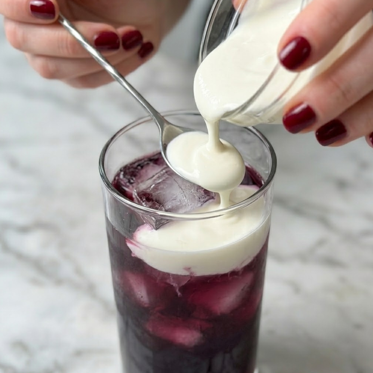 Tight zoom of hands with red nails pouring white cream over the back of a spoon into a purple cocktail.