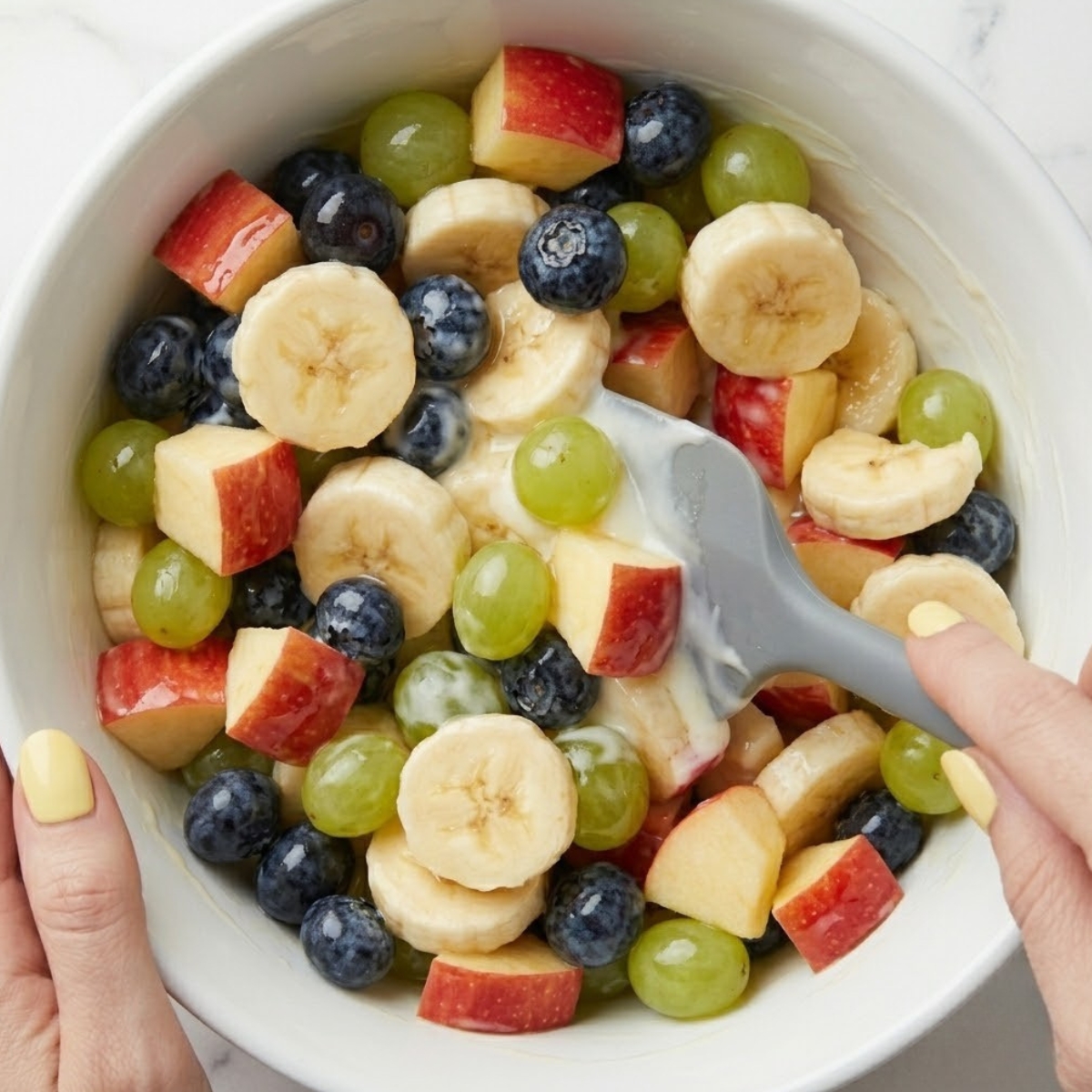 Tight zoom of hands with yellow nails using a silicone spatula to gently toss mixed fresh fruits with creamy yogurt dressing.