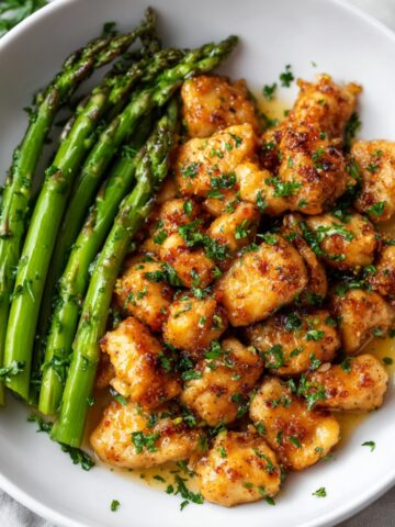 A white bowl of Garlic Butter Chicken bites with sautéed asparagus, coated in a golden garlic butter sauce and garnished with fresh chopped parsley.