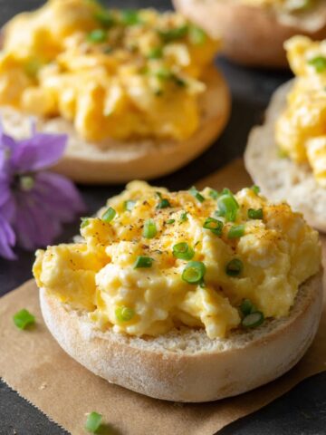 "Fluffy Garlic Herb Boursin scrambled eggs piled on toasted English muffin halves, garnished with sliced green onions and black pepper, with a purple flower beside them."2 / 2