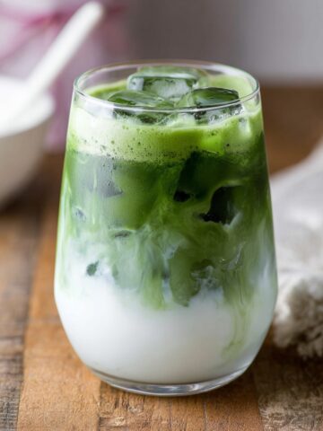 A glass of Iced Coconut Matcha Latte with vibrant green matcha swirling into creamy white coconut milk over ice, served on a wooden board.