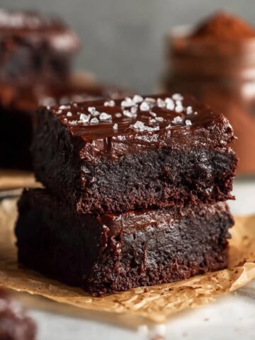 Two stacked Keto Avocado Brownies with a glossy chocolate glaze and flaky sea salt on top, placed on parchment paper with a jar of cocoa powder in the background.