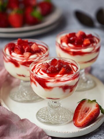 Three elegant crystal dessert glasses filled with creamy white mascarpone mousse swirled with vibrant red strawberry sauce and topped with fresh diced strawberries, arranged on a round white ceramic plate. A halved fresh strawberry sits beside the glasses, with a bowl of whole strawberries and dark chocolate pieces visible in the soft-focus background, alongside a dusty pink linen napkin on a gray surface.