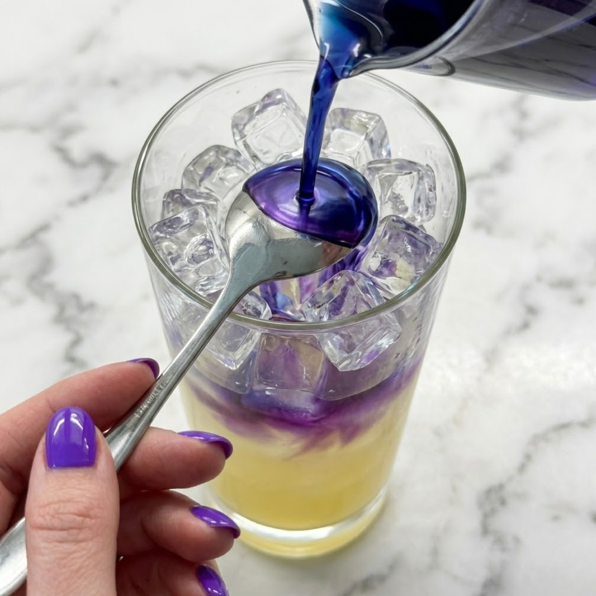 Tight zoom of hands with purple nails pouring blue tea over the back of a spoon into lemonade to create a color-changing effect.