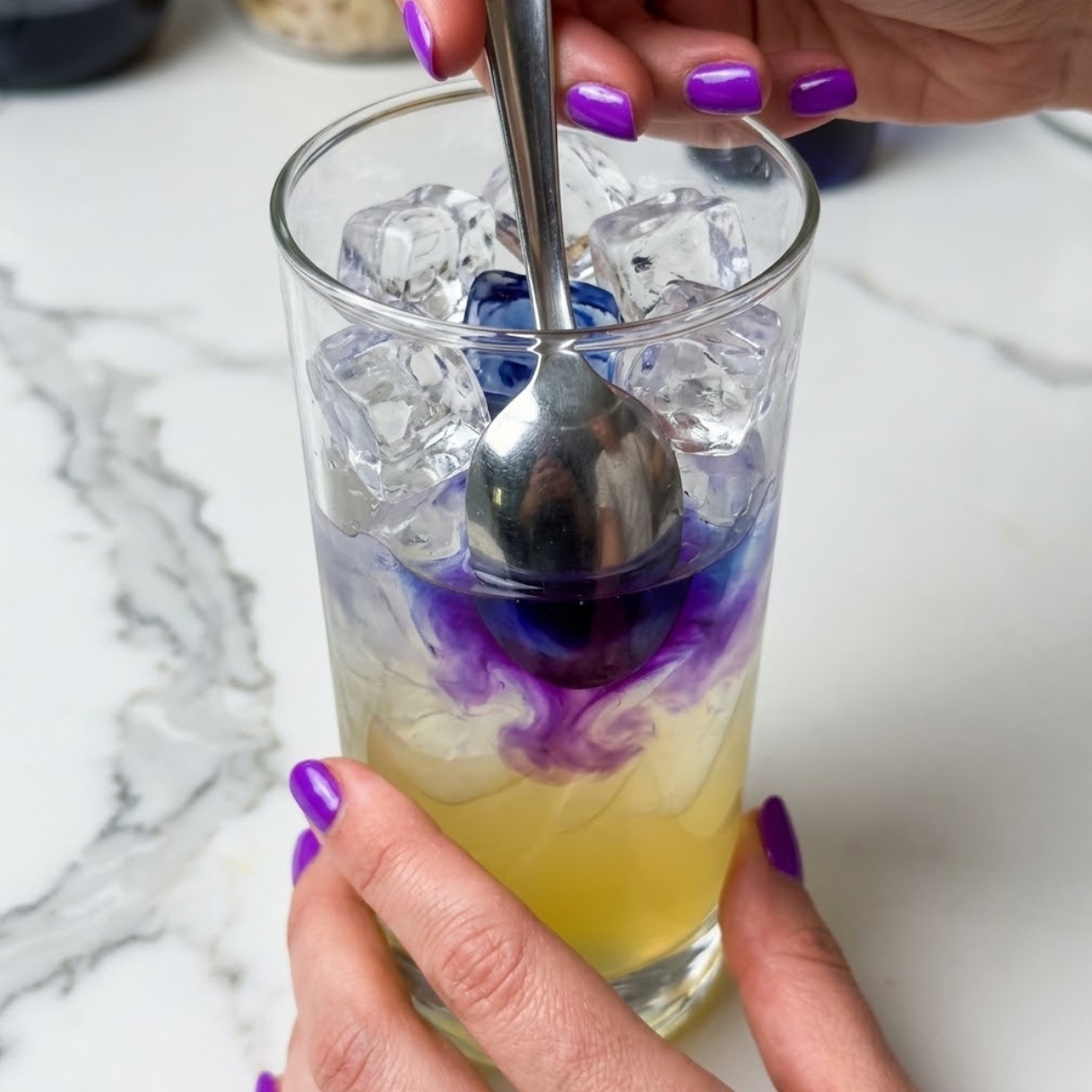 Tight zoom of hands with purple nails pouring blue tea over the back of a spoon into lemonade to create a color-changing effect.