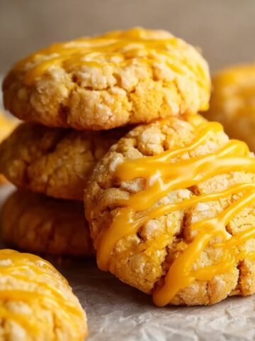 Close-up of golden mango cookies with a crinkled sugar-crusted texture, drizzled with vibrant bright yellow mango glaze in diagonal lines, stacked and scattered on crinkled parchment paper on a light gray surface, with additional cookies softly blurred in the warm background.