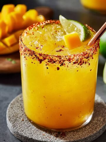 A rocks glass filled with a vibrant golden mango margarita with a spicy red chili and salt rimmed edge, garnished with a fresh lime wedge, a cube of fresh mango, and a rose gold metal straw, placed on a round stone coaster on a dark gray surface, with fresh diced mango on a wooden board and halved limes softly blurred in the sunny background.