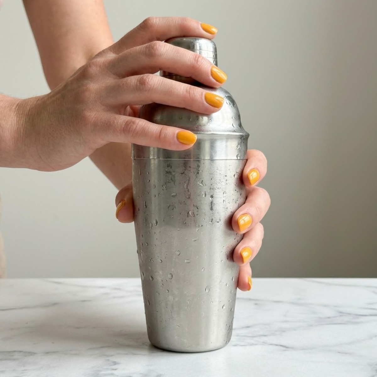 Extreme close-up of hands with yellow nails shaking a cold metal cocktail shaker.